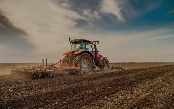 Rode tractor ploegt boerenveld onder bewolkte hemel, land voorbereidend voor magazijnbouwproject. 
